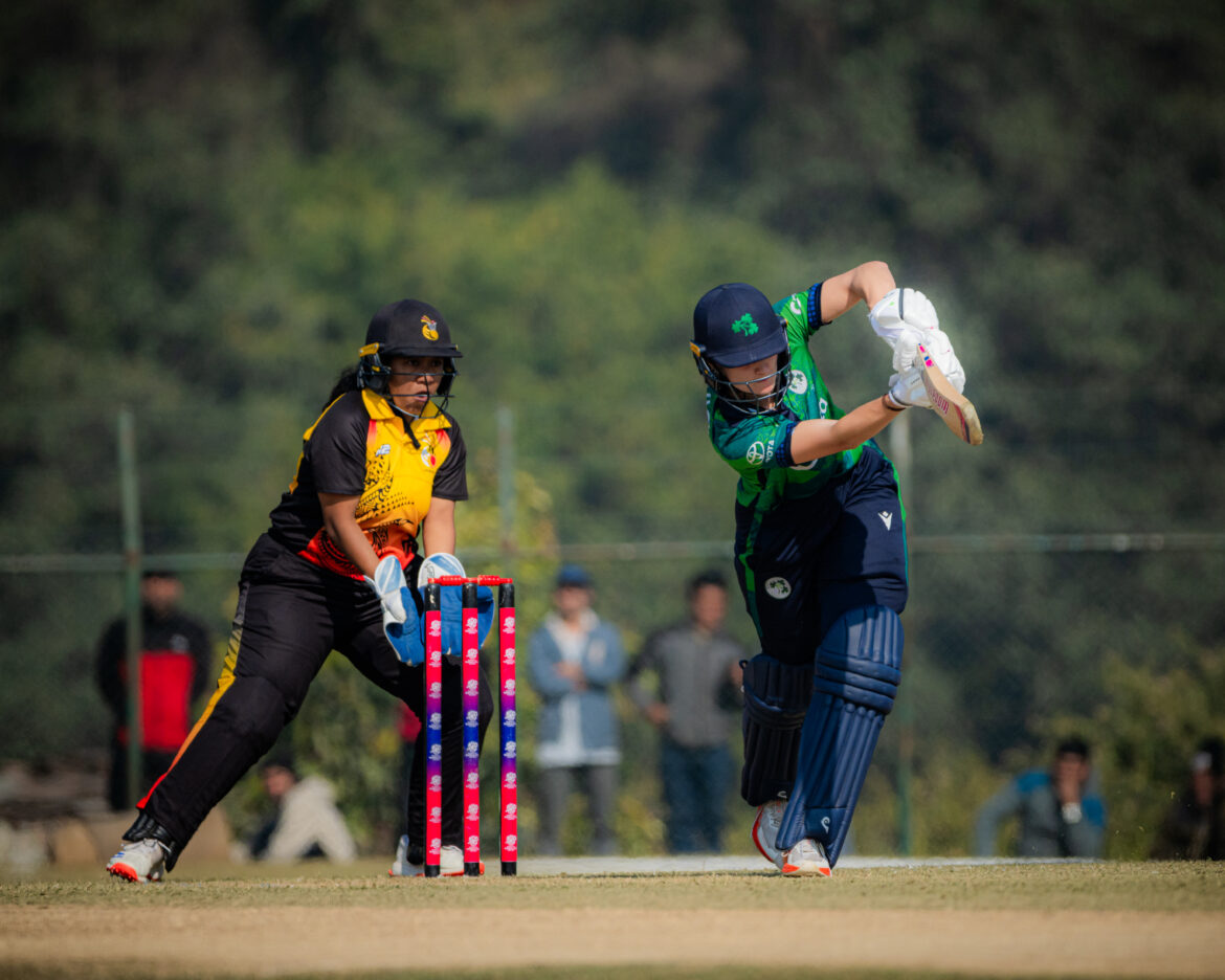 Two women in motion playing cricket. One in a green shirt holding a bat and another wearing a red, black and gold shirt standing behind the wicket.