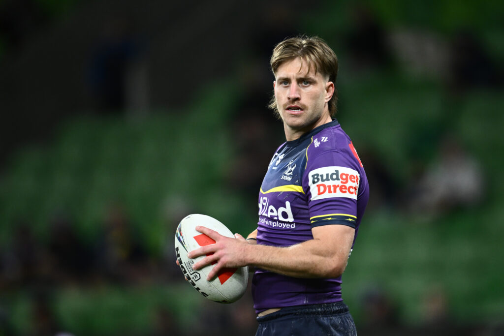 Blond haired caucasian Melbourne Storm player, Cameron Munster, wearing a purple rugbu jersey and holding a rugby ball.