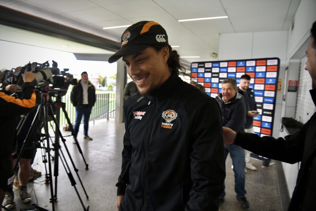Jarome Luai walking away with a smile on his face and wearing a black cap and black jacket with the orange, white and black Wests Tigers Logo on it