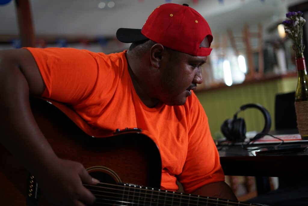 A bulky guy playing the guitar wearing a bright orange shirt.