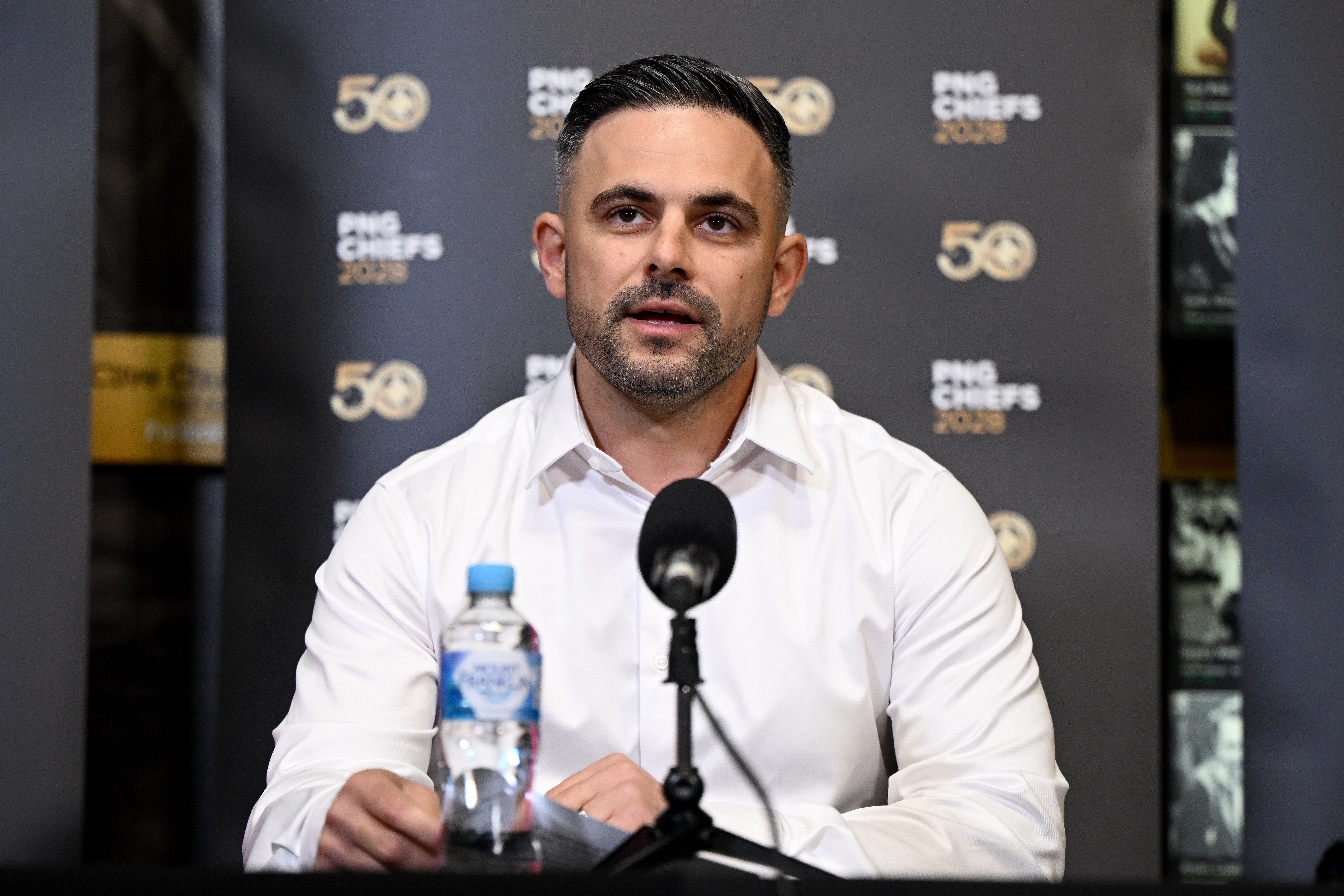 Caucasian man wearing a white button up shirt sitting in front of a microphone at a press conference.