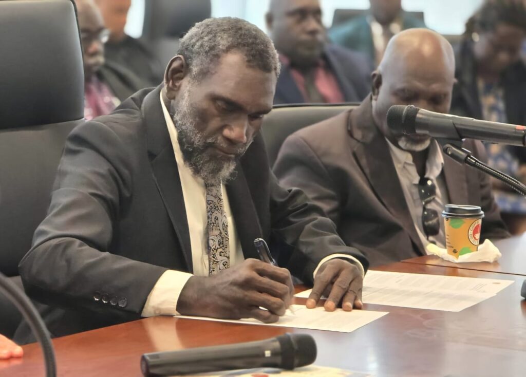 Dark skinned man in a suit signing a document on a brown table.